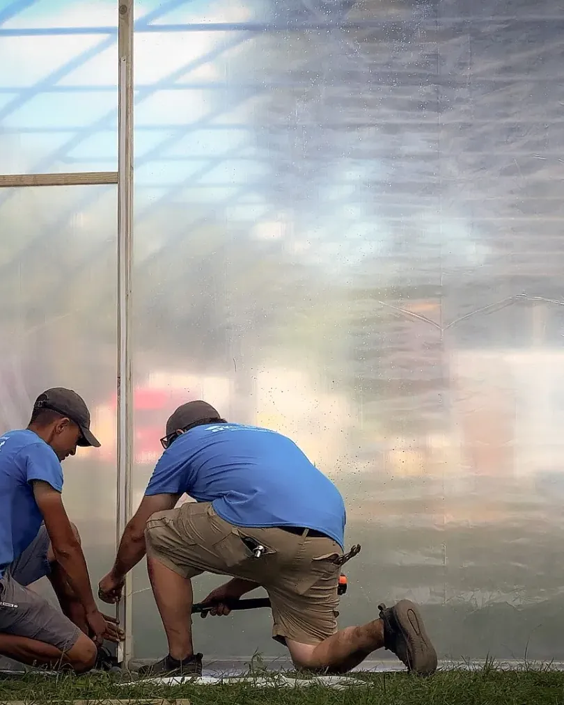 Two men kneeling side by side, working together at the base of a greenhouse wall.